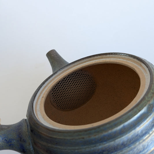 Close-up of a ceramic teapot with a blue-gray glazed exterior, a brown interior, and a metal mesh infuser visible inside the open top, set against a plain light background.