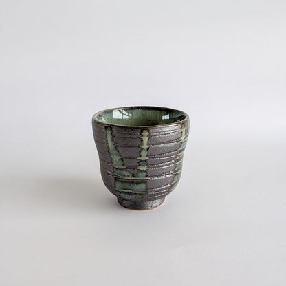 A handmade ceramic tea cup with a textured, ribbed exterior in dark gray and green glaze, set against a plain white background.